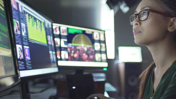 Close up stock image of a young asian woman sitting down at her desk where she’s surrounded by 3 large computer monitors displaying out of focus images of people as thumbnails; crowds; graphs &amp; scrolling text.